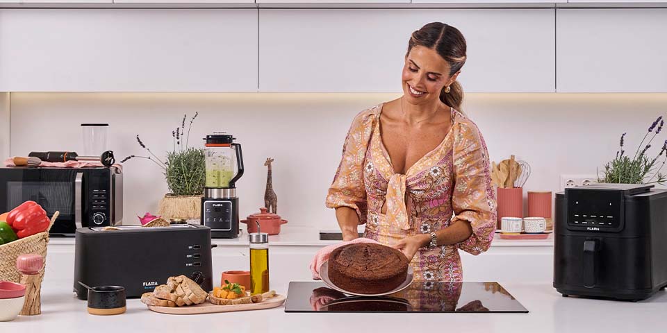 Catarina Gouveia na cozinha a segurar um bolo. À sua volta, eletrodomésticos Flama - torradeira, micro-ondas, liquidificadora e fritadeira sem óleo Air Fryer