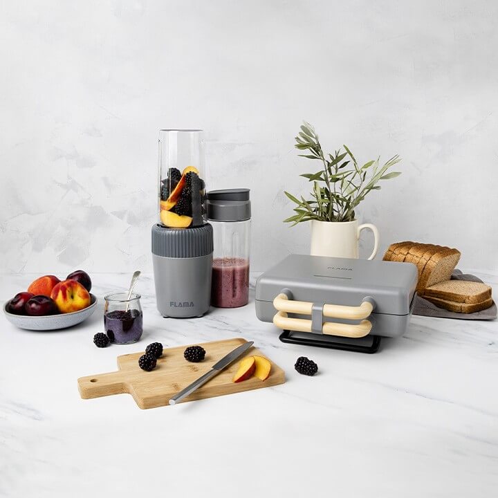 Conjunto de cozinha com uma sanduicheira e uma liquidificadora acompanhada de tostas, framboesas, pêssegos e outras frutas com cores vibrantes.